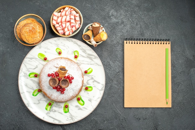 Foto gratuita vista dall'alto da lontano una torta una torta appetitosa con i frutti di bosco biscotti caramelle cialde notebook matita