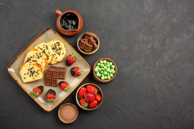 Top view from afar cake and strawberries bowls of chocolate strawberries green candies and chocolate cream next to the plate of cake with chocolate-covered strawberries