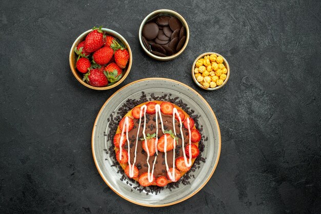 ストロベリーヘーゼルナッツとチョコレートのチョコレートボウルと暗いテーブルにチョコレートとイチゴのケーキと遠くのケーキからの上面図