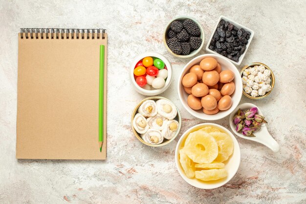 Top view from afar candies in bowls appetizing candies and dried pineapples in bowls next to the cream notebook and green pencil