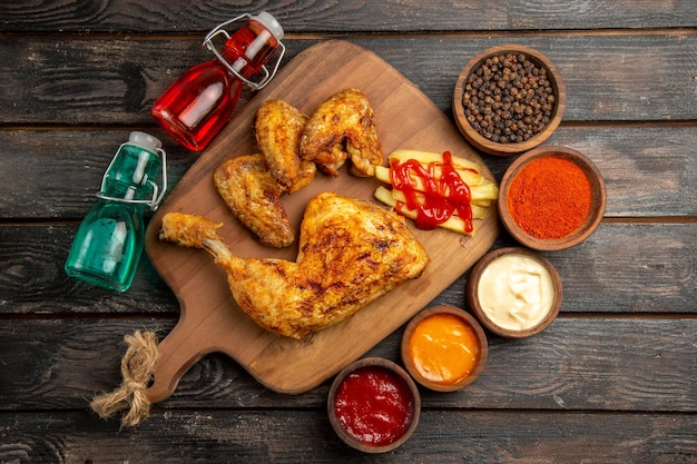 Top view from afar chicken red and blue bottles next to the chicken with french fries and ketchup on the cutting board bowls of black pepper sauces spices