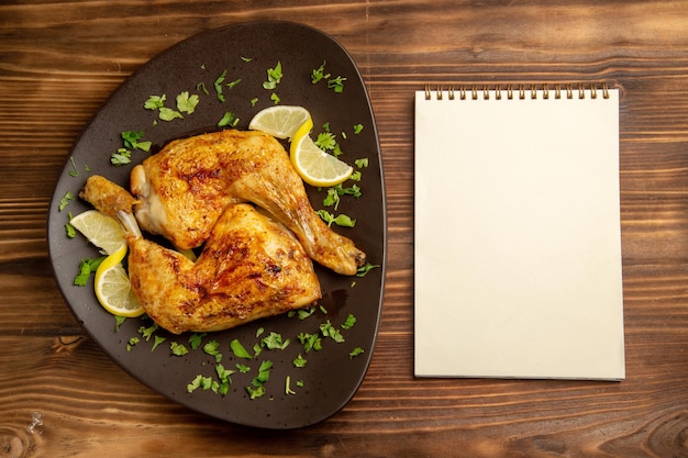 Foto gratuita vista dall'alto da lontano pollo con taccuino bianco limone accanto al piatto di un appetitoso pollo con erbe e limone sul tavolo di legno