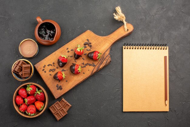 Top view from afar chocolate strawberries chocolate cream and strawberries in bowls and chocolate-covered strawberries on the kitchen cutting board next to notebook and pencil