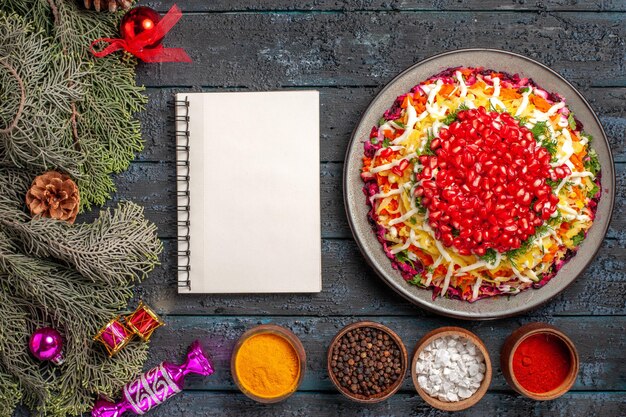 Top view from afar Christmas food Christmas dish with pomegranate bowls of spices white notebook and spruce branches with Christmas tree toys