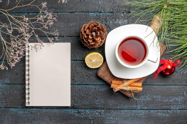 Top view from afar Christmas tree branches a cup of black tea cinnamon sticks on the wooden board next to the Christmas spruce branches tree toys white notebook and lemon