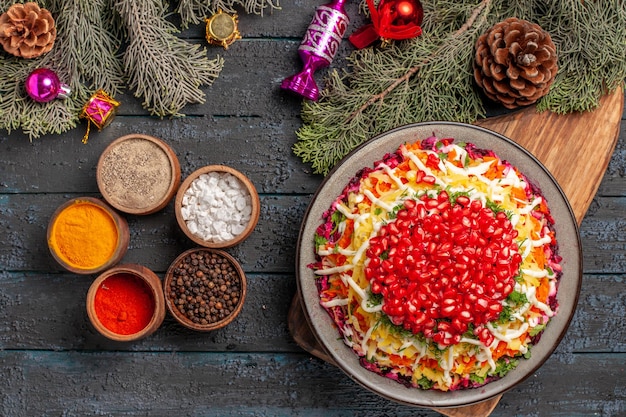 Top view from afar Christmas tree toys dish with pomegranates on the cutting board bowls of spices on the dark surface next to the tree branches with cones and Christmas tree toys