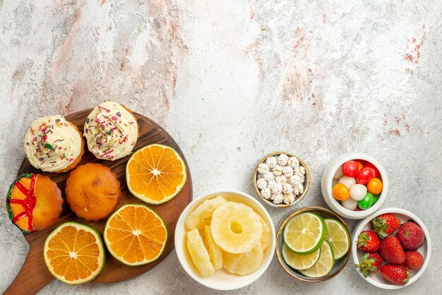 Top view from afar citrus fruit on the board bowls of strawberries limes sweets and dried pineapples and sliced orange and cookies on the wooden cutting board