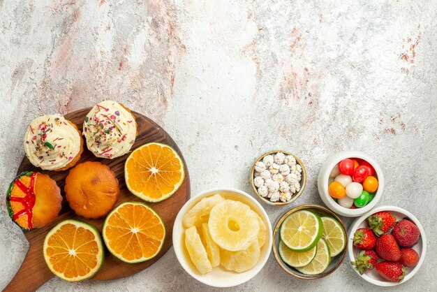 Вид сверху цитрусовых на доске, миски, клубника, лайм, сладости и сушеные ананасы, нарезанный апельсин и печенье на деревянной разделочной доске