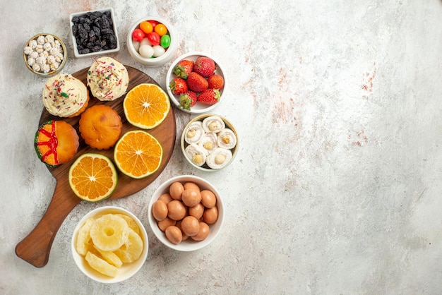 Top view from afar citrus fruit on the board sliced orange and cookies on the wooden cutting board next to the bowls of sweets and dried pineapples