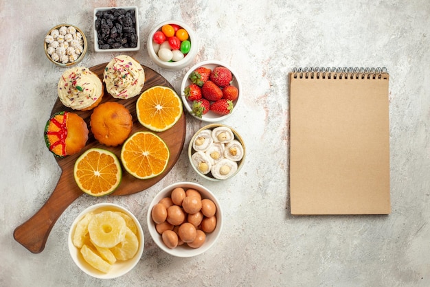 Top view from afar citrus fruit on the board sliced orange and cookies on the wooden cutting board next to the cream notebook bowls of sweets and dried pineapples