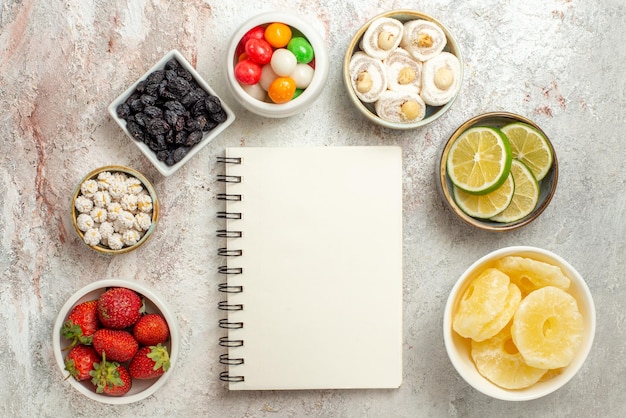 Top view from afar citrus fruits bowls of berries sweets and dried pineapples are laid out in a circle next to the white notebook