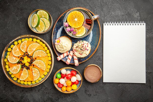 Top view from afar a cup of herbal tea cupcakes with cream and the cup of herbal tea on the blue saucer next to the white notebook and bowls of citrus fruits chocolate cream and candies on the table