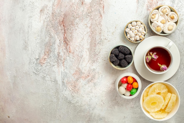 Top view from afar cup of tea a cup of tea and bowls of different sweets dried pineapples Turkish delight on the white background