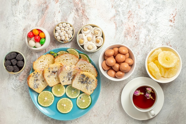 Top view from afar cup of tea white cup of tea and bowls of appetizing Turkish delight and dried pineapples next to the plate of cake and sliced limes on the table