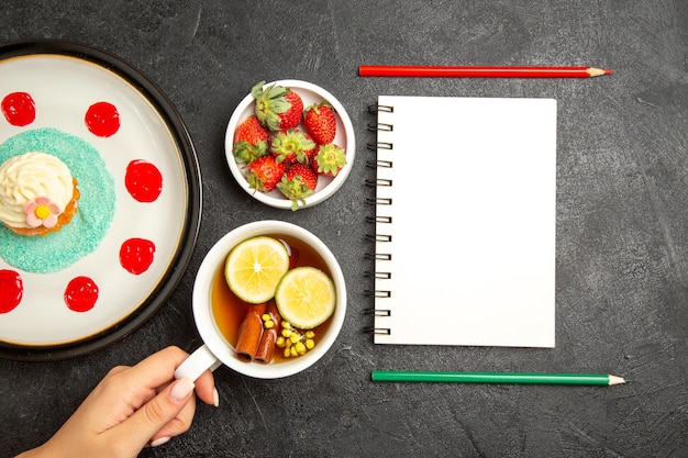 Vista dall'alto da lontano cupcake una tazza di tè piatto di cupcake ciotola di fragole accanto al quaderno bianco con due matite e la tazza di tè con limone in mano