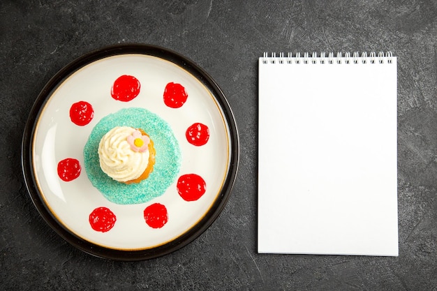 Top view from afar cupcake on the plate white notebook next to the cupcake with sauce on the white plate on the dark table