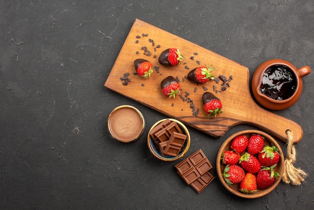 Top view from afar cutting board chocolate-covered strawberries on cutting board next to chocolate cream and strawberries on the right side of the dark table