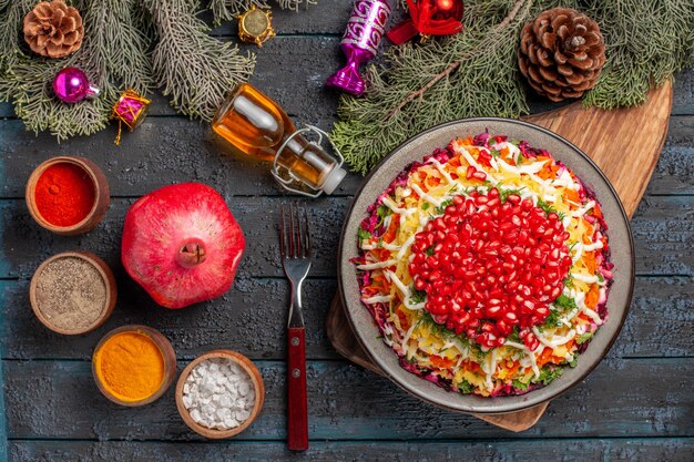 Top view from afar dish in the plate dish with pomegranate on the board next to the bowls of spices bottle of oil fork pomegranate and tree branches with cones and Christmas tree toys