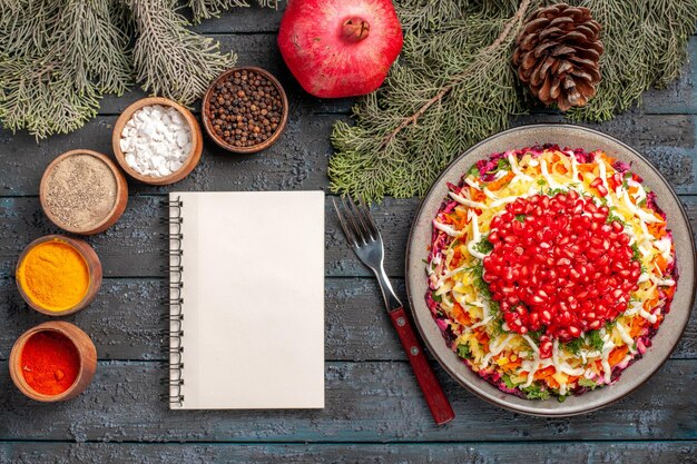 Top view from afar dish and tree branches dish of pomegranate potatoes in white plate next to the white notebook bowls of spices fork pomegranate and tree branches with cones on the dark table