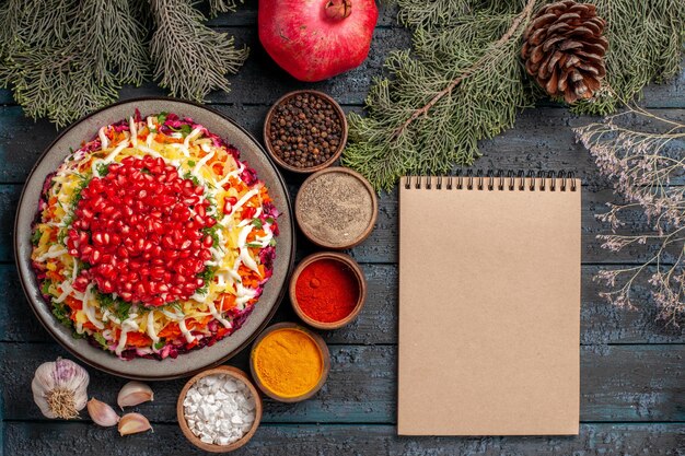 Top view from afar dish and tree branches dish of seeds of pomegranates next to the bowls of spices pomegranate cream notebook and tree branches cones on the grey table