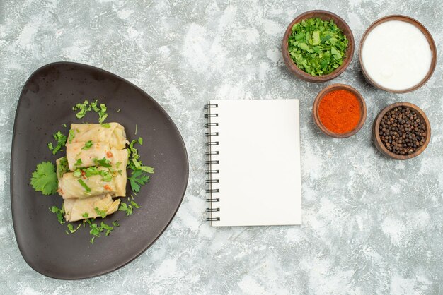 Top view from afar dish with herbs plate of stuffed cabbage next to white notebook and bowls with black pepper sour cream herbs and spices on the left and right side of the table