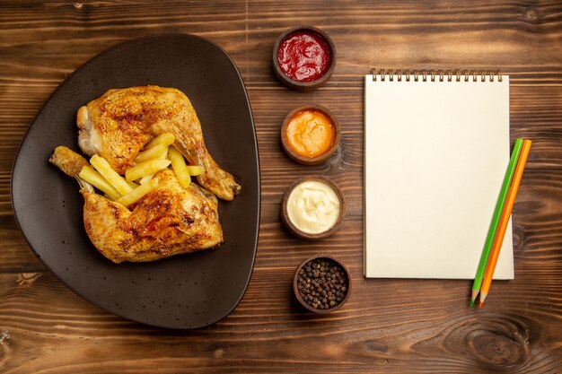 Top view from afar fast food white notebook two pencil bowls of colorful sauces and black pepper next to the plate of chicken and french fries