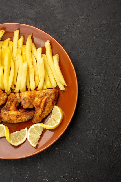 검정 탁자 왼쪽에 있는 식욕을 돋우는 감자튀김 닭 날개와 레몬을 접시에 담은 멀리 있는 음식의 꼭대기