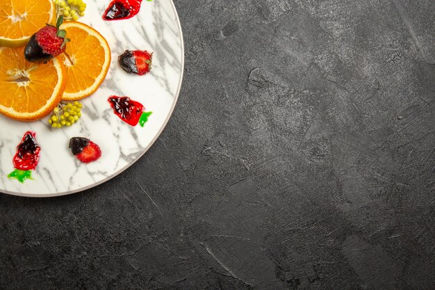 Вид сверху издалека фрукты на тарелке клубника в шоколаде и цитрусовые на белой тарелке на темном столе