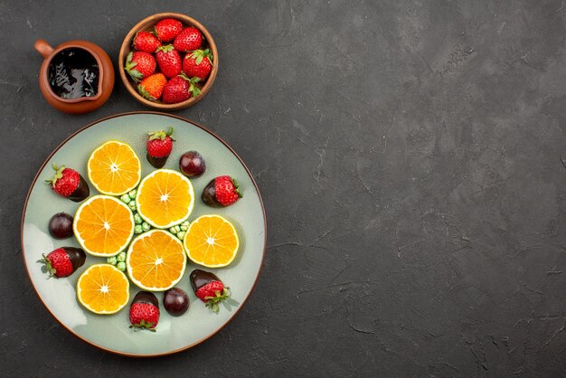 Top view from afar orange and chocolate bowls of chocolate sauce and strawberries next to plate of chocolate-covered strawberry chopped orange green candies on the left side of the dark table