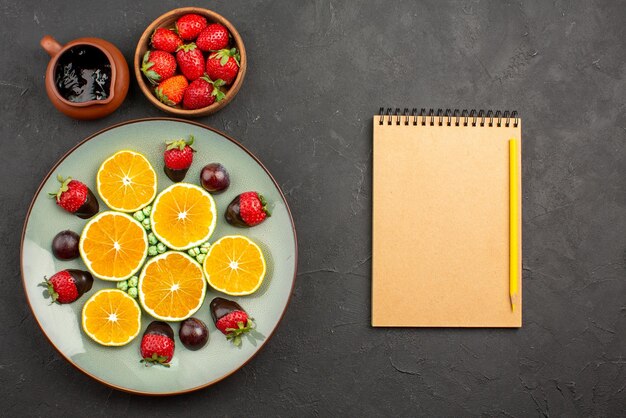 Top view from afar orange and chocolate chocolate sauce and strawberries next to chocolate-covered strawberry chopped orange green candies and notebook with pencil