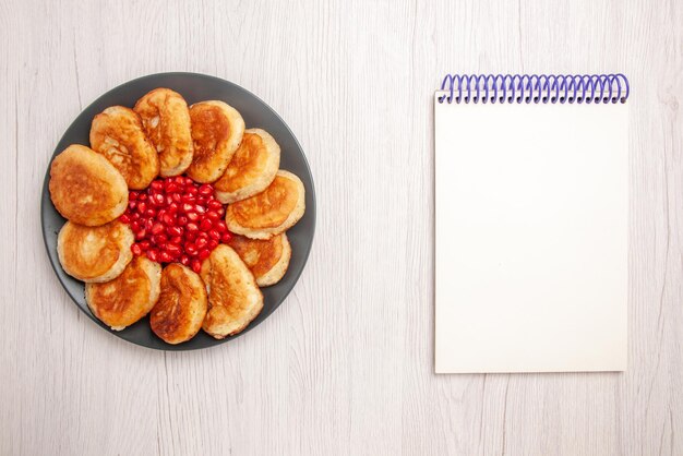 白いテーブルの上の白いノートの横にある黒いプレートにザクロのパンケーキを食欲をそそる遠くのパンケーキからの上面図