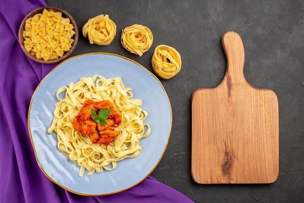 Foto gratuita vista dall'alto da lontano ciotole di pasta e carne di pasta e piatto di sugo di pasta e carne sulla tovaglia viola accanto al tagliere