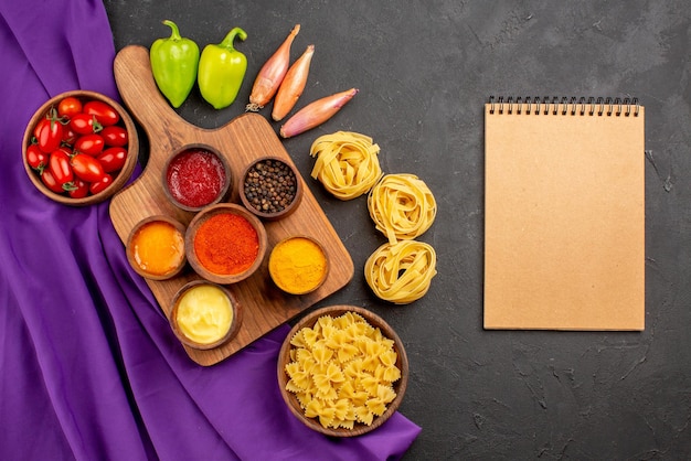 Top view from afar pasta and tomatoes spices and sauces in bowls on the cutting board onion bowl of tomatoes ball pepper and pasta next to the cream notebook on the table