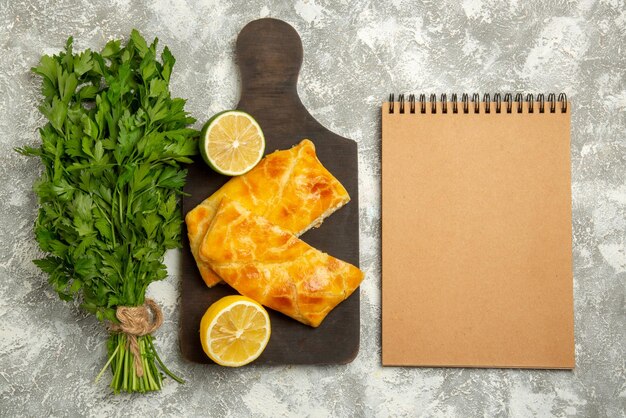 Top view from afar pies herbs cheese pies and lemon on the wooden board next to the cream notebook on the table