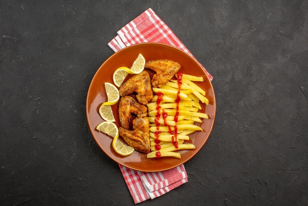 Top view from afar plate on tablecloth orange plate of chicken wings ketchup french fries and pieces of lemon on pink-white checkered tablecloth in the center of the dark table