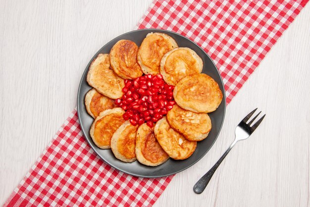 Top view from afar pomegranate appetizing pancakes and seeds of pomegranate on the checkered tablecloth and a fork on the table