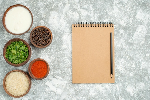 Top view from afar rice and spices bowl of rice herbs sour cream spices and black pepper next to cream notebook and pencil on the left side of the table