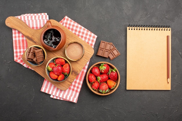 Top view from afar strawberries and chocolate strawberries chocolate cream in bowl on the cutting board on tablecloth next to the plate of strawberries and notebook with pencil