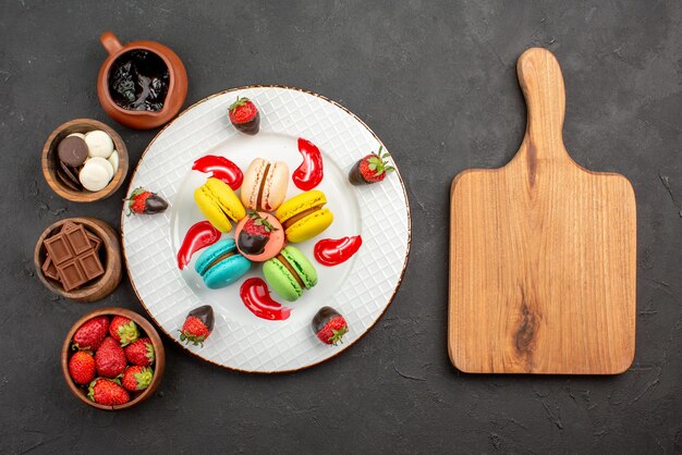 Top view from afar sweet dish plate of macaroon and four bowls of candies chocolate strawberries and chocolate cream next to the wooden cutting board on the dark background