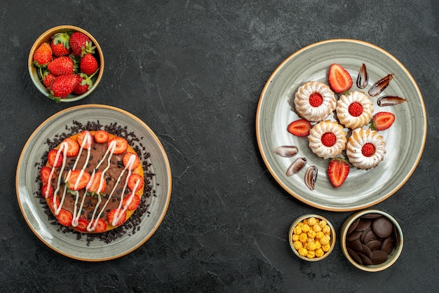 Vista dall'alto da lontano dolci e torta appetitosa torta e biscotti con fragola e cioccolato e ciotole di nocciole al cioccolato e fragola sul tavolo nero