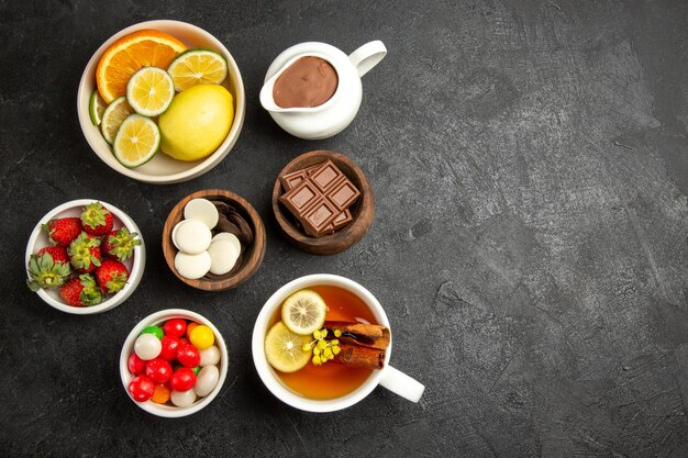 Вид сверху издалека сладости на столе чашка чая с корицей и лимоном и миски клубники, шоколада и конфет на темном столе