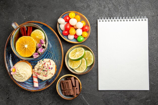 Top view from afar sweets on the table three bowls of candies chocolate and slices of limes next to the white notebook two cupcakes and the cup of herbal tea on the table