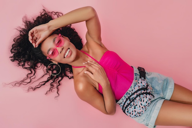 Top view from above attractive black african american woman lying in stylish summer outfit pink top shorts on pink background summer fashion trend happy smiling curly hair sunglasses accessories