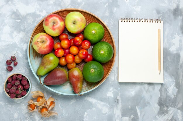 Top view fruit composition apples pears tangerines and plums notepad on white desk.