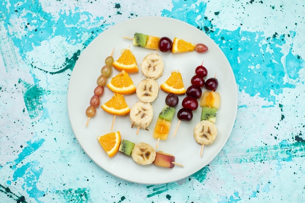 Вид сверху фруктовой композиции, нарезанной на палочках внутри белой тарелки на ярком столе, фруктового экзотического печенья, сахара