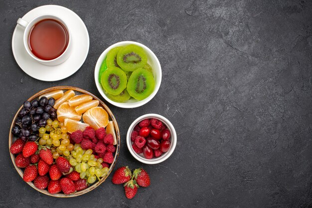 Top view fruit composition strawberries grapes raspberries and tangerines with cup of tea on dark-grey desk