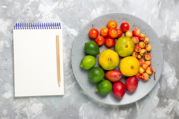 Top view fruit composition tangerines lemons plums inside plate on light grey desk.