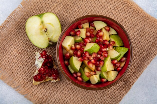 Top view of fruit salad including pomegranate seeds and chopped apples in a red bowl with half green apple and pomegranate slice on sack cloth surface