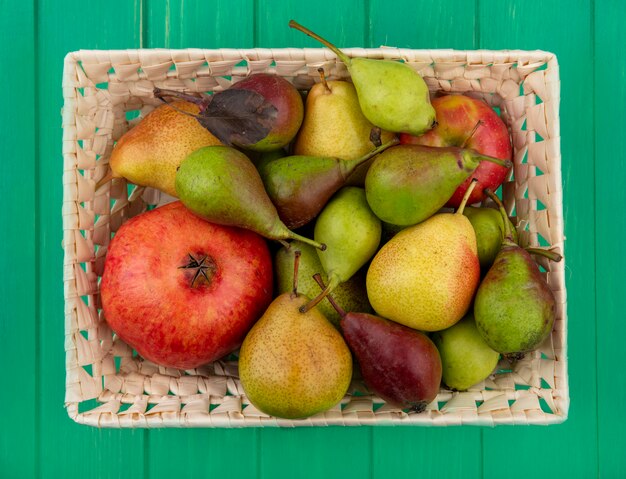 Вид сверху на фрукты, такие как яблоко, гранат, груши и персик в корзине на зеленой поверхности