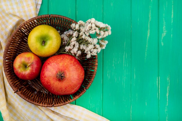 Вид сверху на фрукты, такие как яблоко и гранат с цветами в корзине на клетчатой ткани и зеленой поверхности